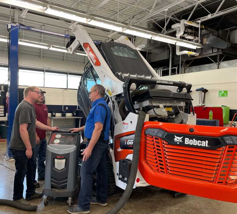 A male instructor and two male students standing in a shop next to a Bobcat