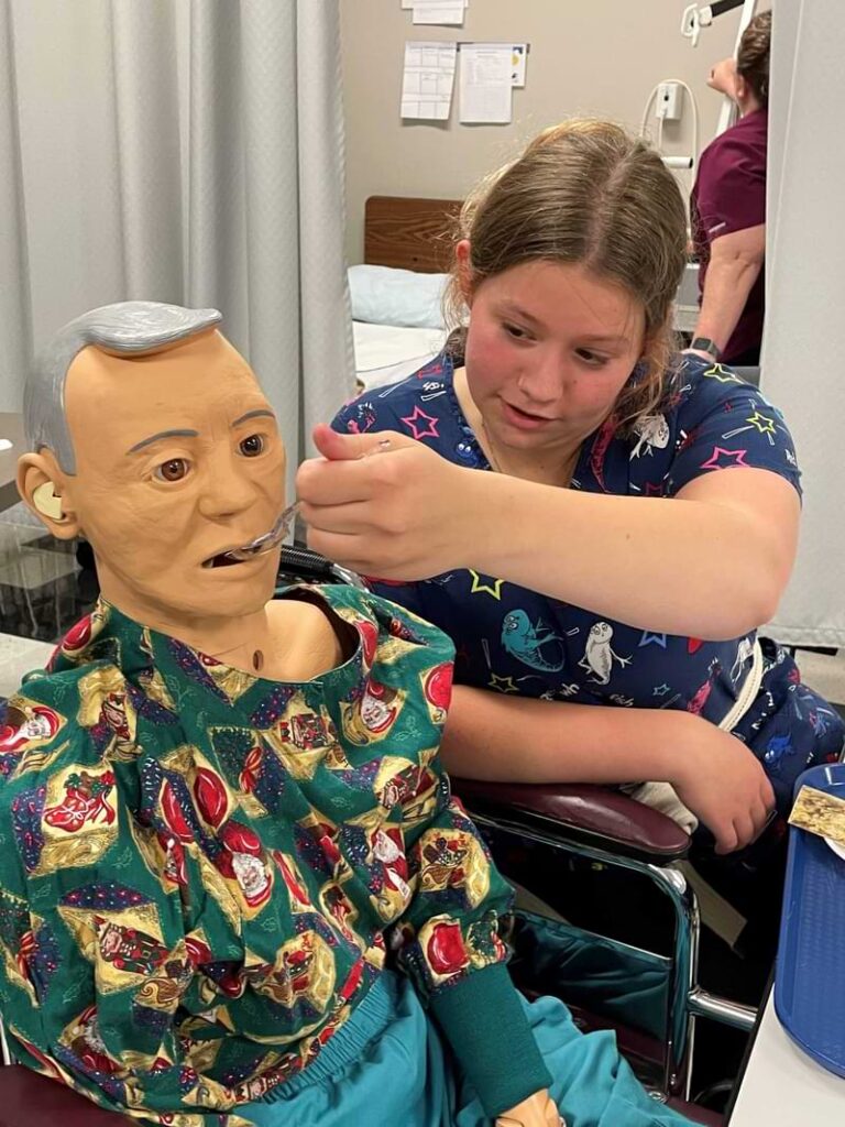 A female student in scrubs feeding a patient (doll)